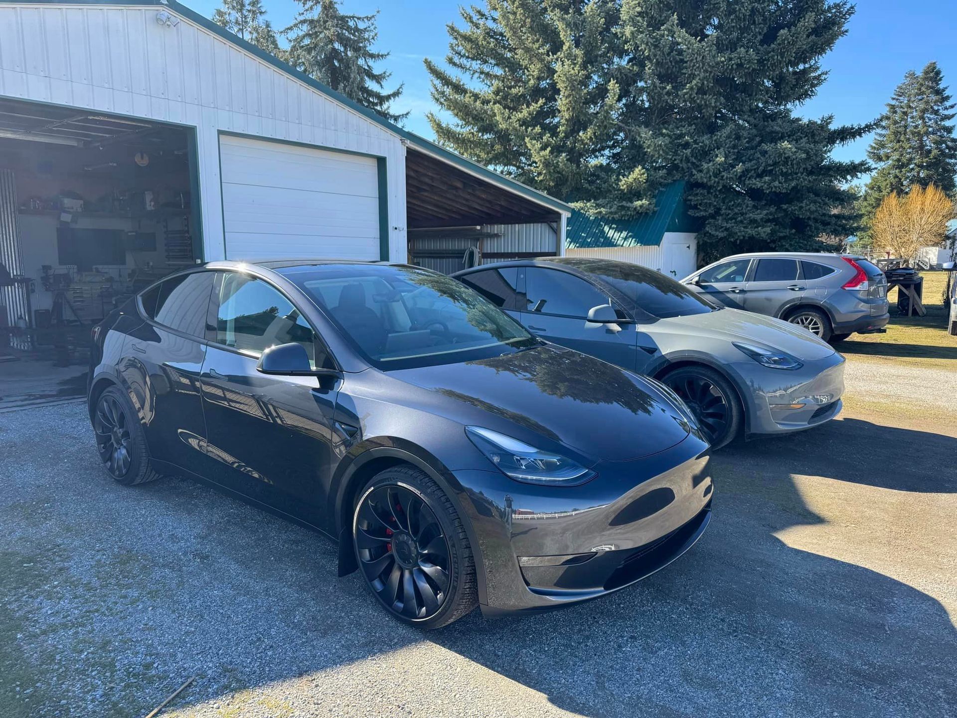 Two Tesla Model Y cars parked in front of a building. One is dark gray, the other silver. Sunny outdoor setting.