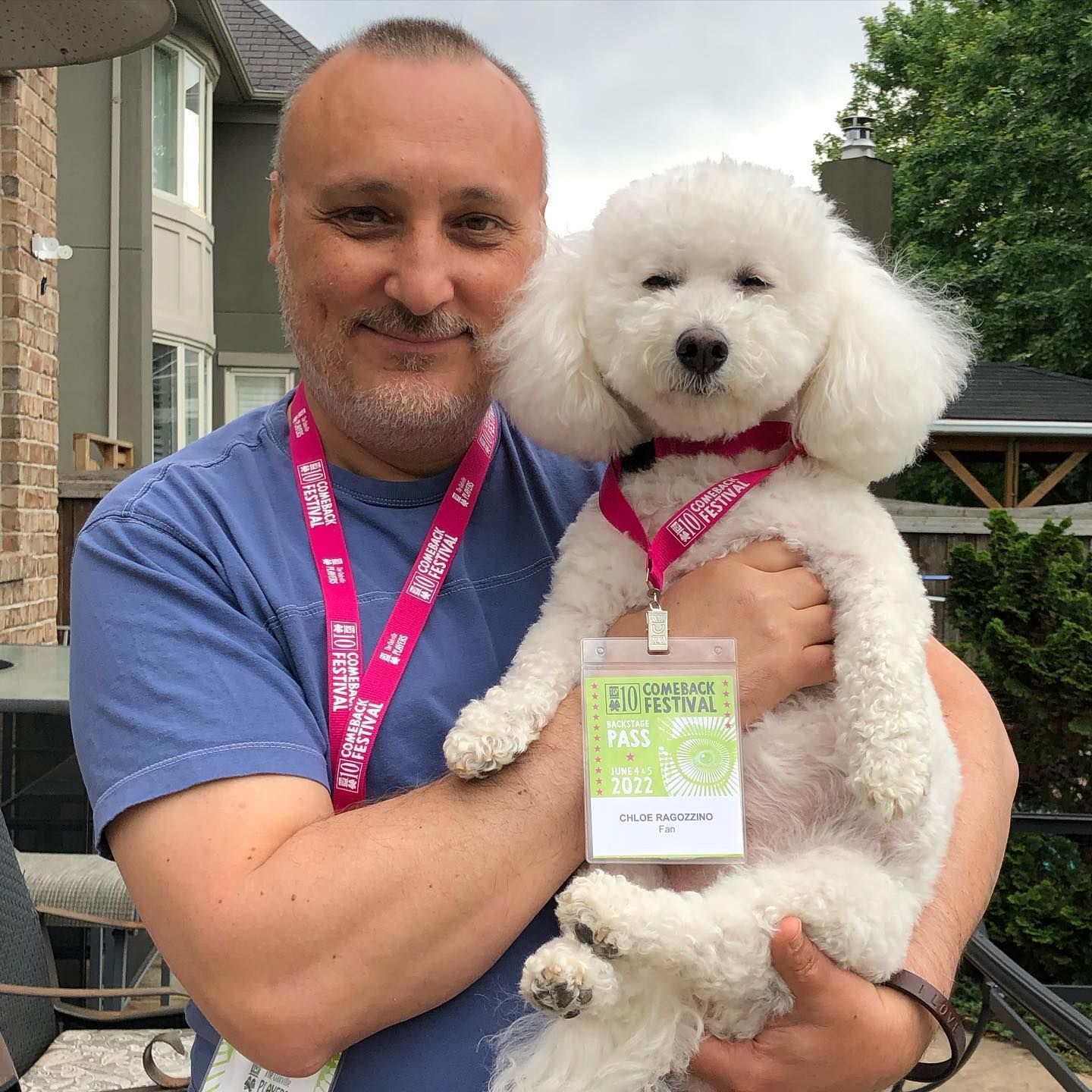Man holding a white fluffy dog wearing a pink lanyard and badge outdoors