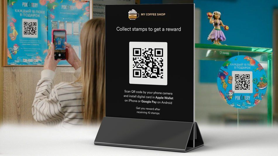 Black tabletop sign with QR code on a desk, with a woman photographing it and colorful posters in the background