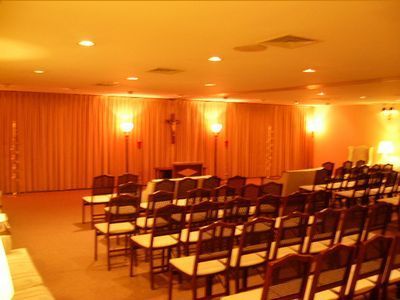 Orange-lit empty room with rows of folding chairs facing a small stage and wall cross