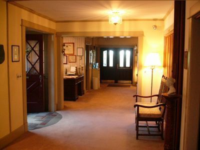 Warmly lit hallway with beige carpet, wood trim, a desk, framed art, and chairs near a lamp