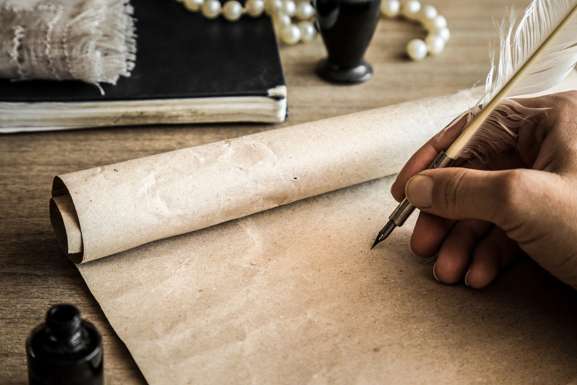Hand writing on scroll with quill pen; inkwell, book, pearls on wood table.