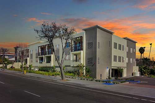 Modern apartment building on a street corner at sunset, with palm trees and colorful sky.