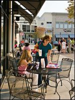 Two people sit at a café table on a sunny sidewalk, with a woman standing beside them and a busy street behind.