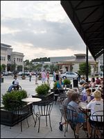 Outdoor café patio with seated people, parked cars, and a city square under an overcast sky