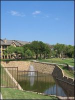 Residential canal with a small fountain, stone walls, green lawns, and houses under a blue sky