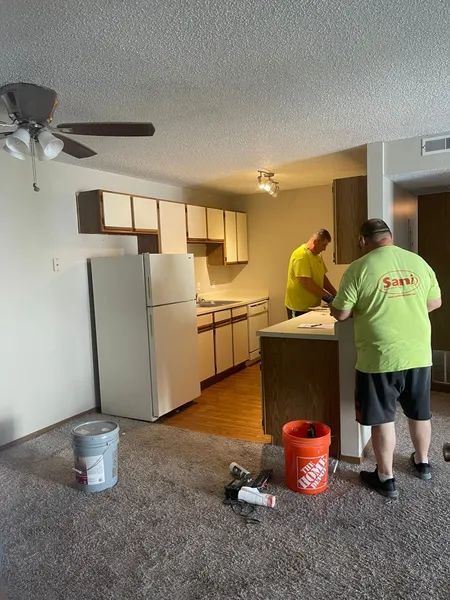 Two people renovating a kitchen, painting cabinets and walls. White refrigerator, brown and white cabinets.