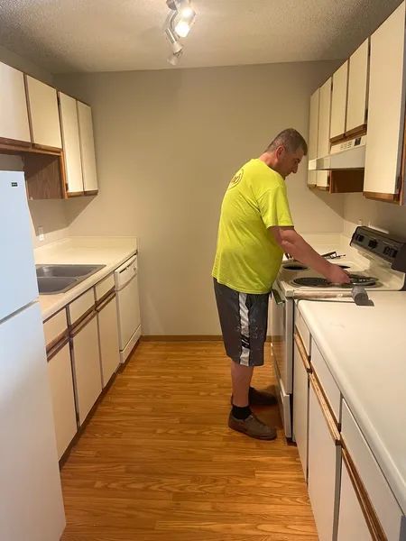 Man in a kitchen with white cabinets and a stovetop. He is wearing a yellow shirt and shorts.