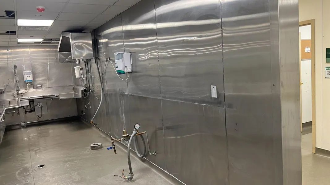Stainless steel industrial kitchen with sinks and spouts; water and debris on the floor and wall.