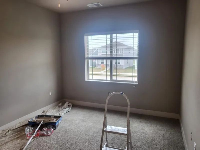 Empty room with painted walls, window, carpet, and painting supplies. A stepladder stands in the room.