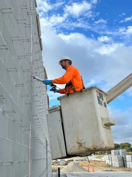 Construction worker in lift, attaching material to a gray block wall under a blue sky.