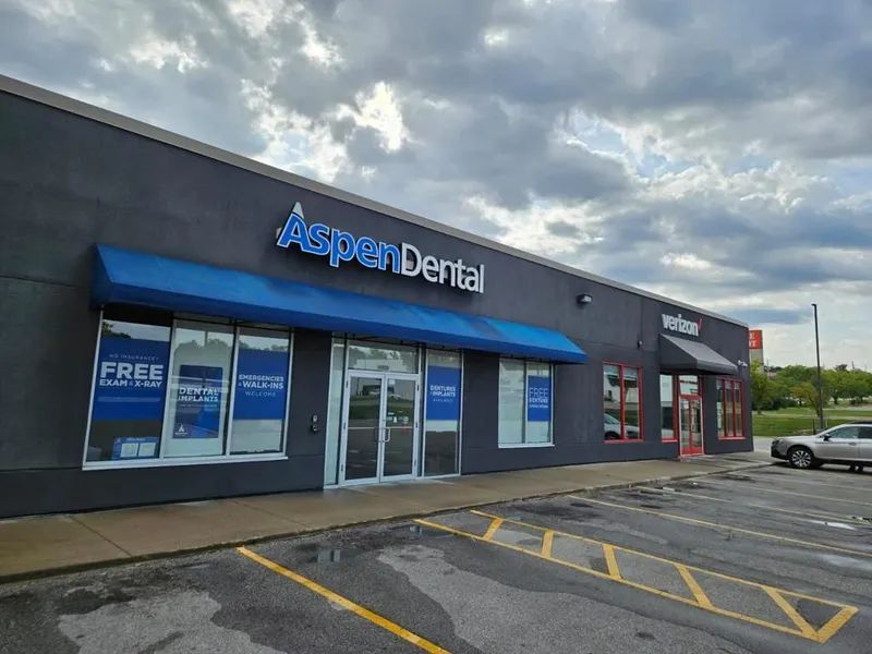 Aspen Dental clinic with blue awning and signage, next to a Verizon store, cloudy sky.