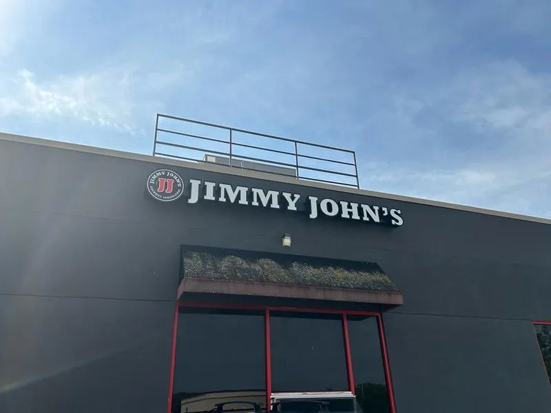 Jimmy John's storefront with white letters and logo on a gray building, under a blue sky.