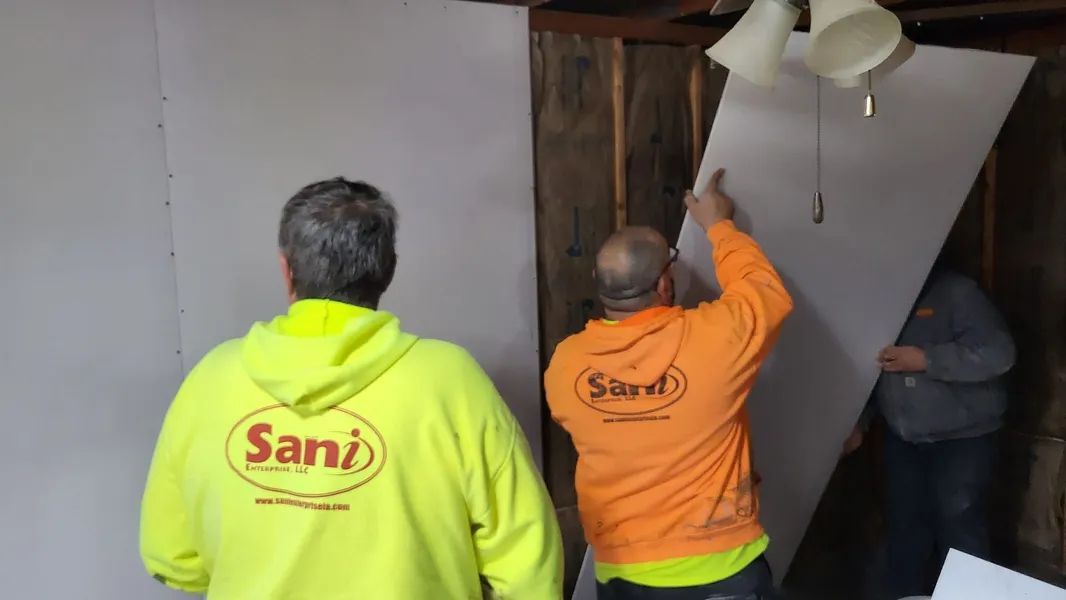Three workers installing drywall in a room. Two wear hoodies with the company logo.