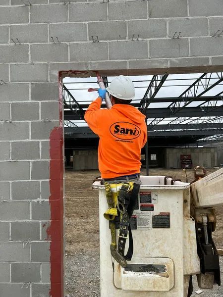 Construction worker in orange hoodie on lift, applying sealant to a concrete block opening.