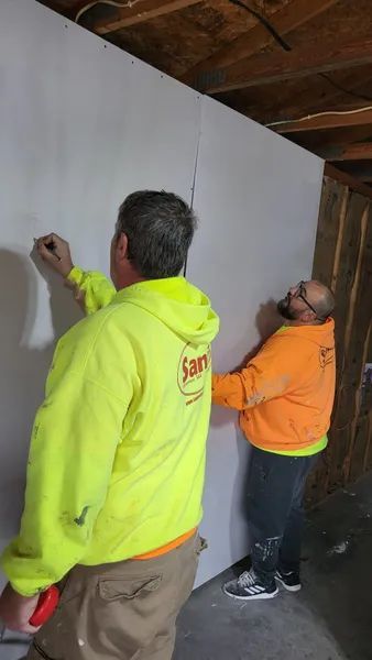 Two people in high-visibility hoodies installing drywall; one draws, the other supports the sheet, indoors.