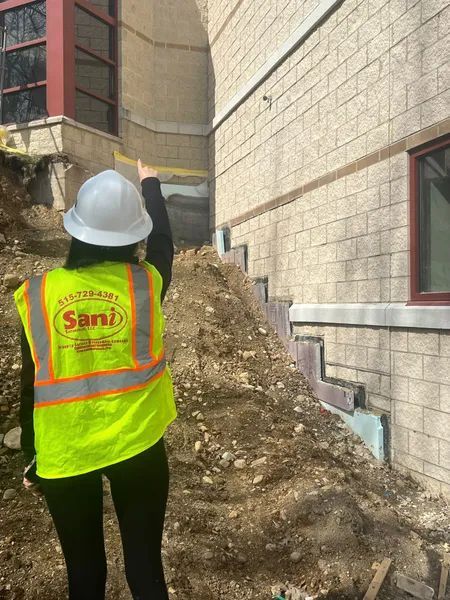 Person in a hard hat and safety vest points at a building's foundation during an outdoor inspection.