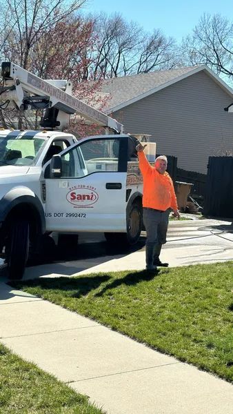 Man in orange shirt waves near a white truck with a lifted boom in a residential setting.