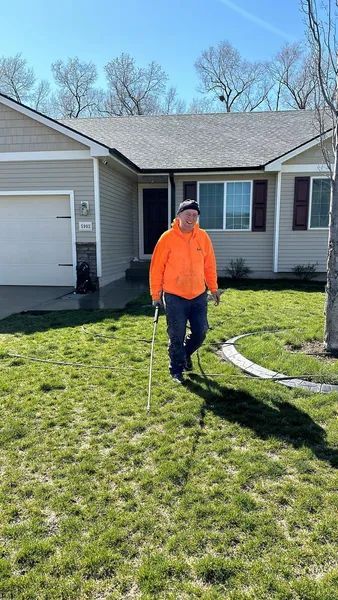 Man in orange jacket walks on lawn in front of a house, holding a line, sunny day.