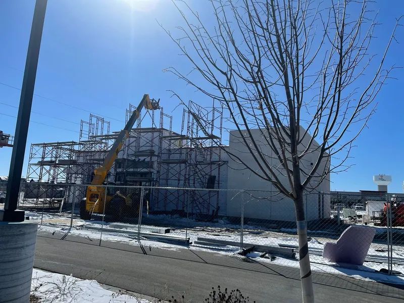 Construction site with yellow crane, metal frame structure, white walls, and a tree against a blue sky.