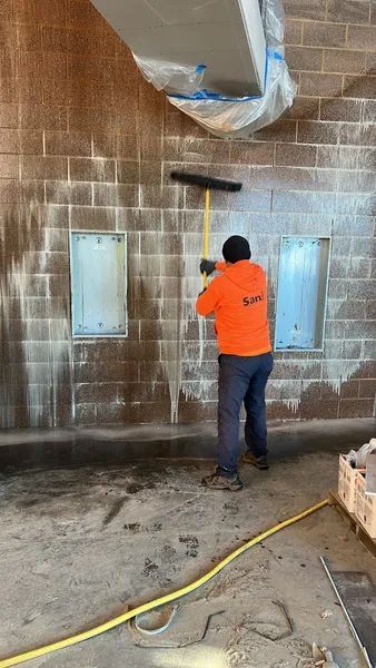 Person in orange shirt cleaning a brick wall with a long-handled brush. Yellow hose on floor.