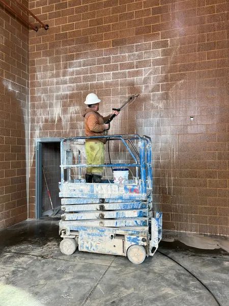 Person on a lift sprays a brick wall indoors; white residue visible.