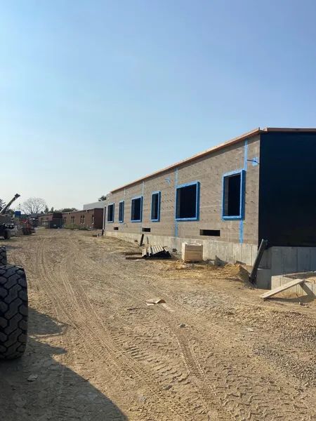 Construction site: Exterior of a long, low building with window frames taped blue. Sunny, dirt ground.