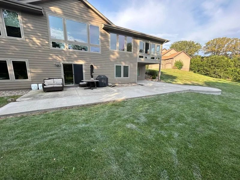 Backyard with concrete patio, grass lawn, and large house with windows.