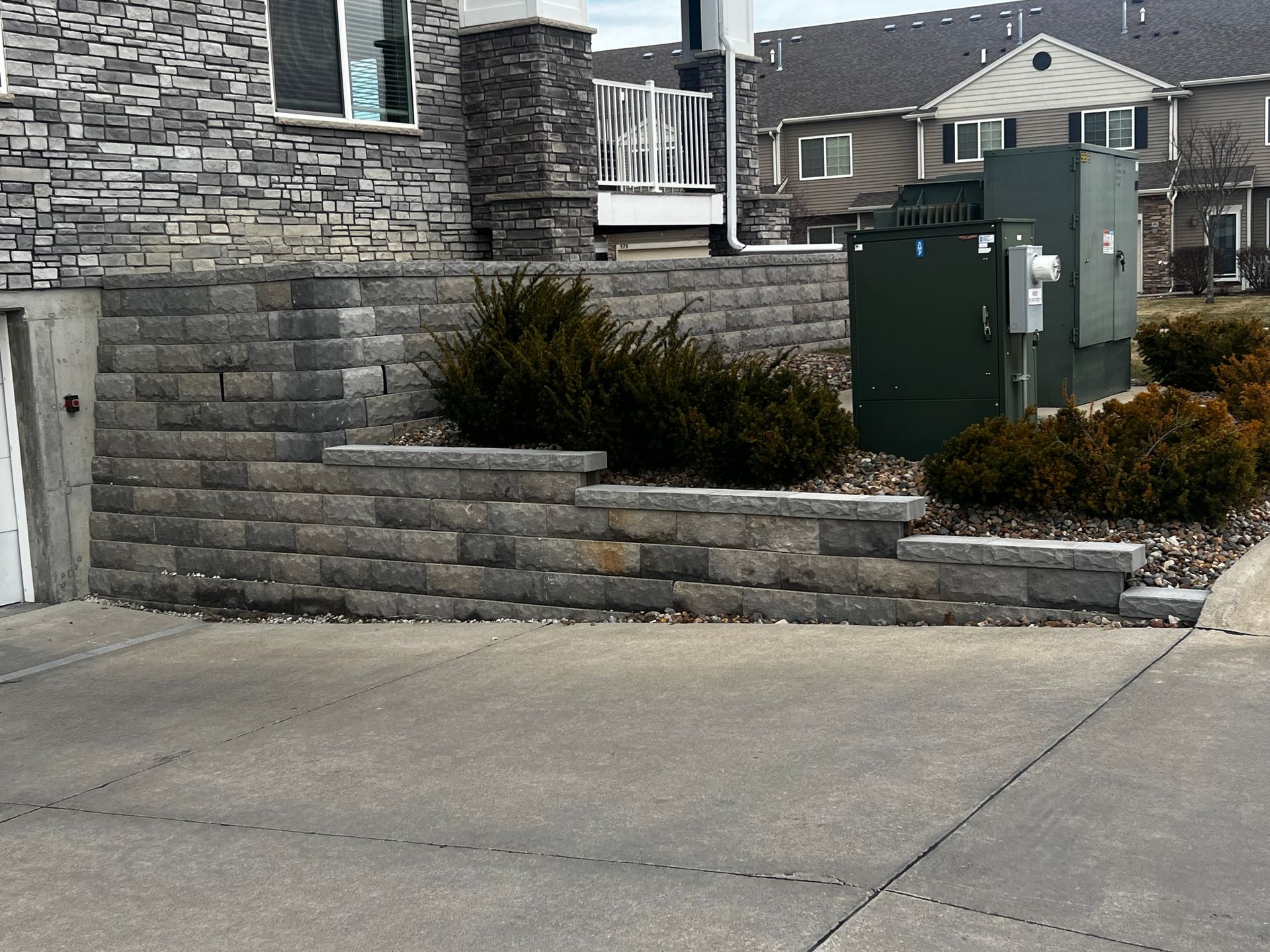 A low retaining wall made of gray bricks borders a concrete driveway and small bushes.