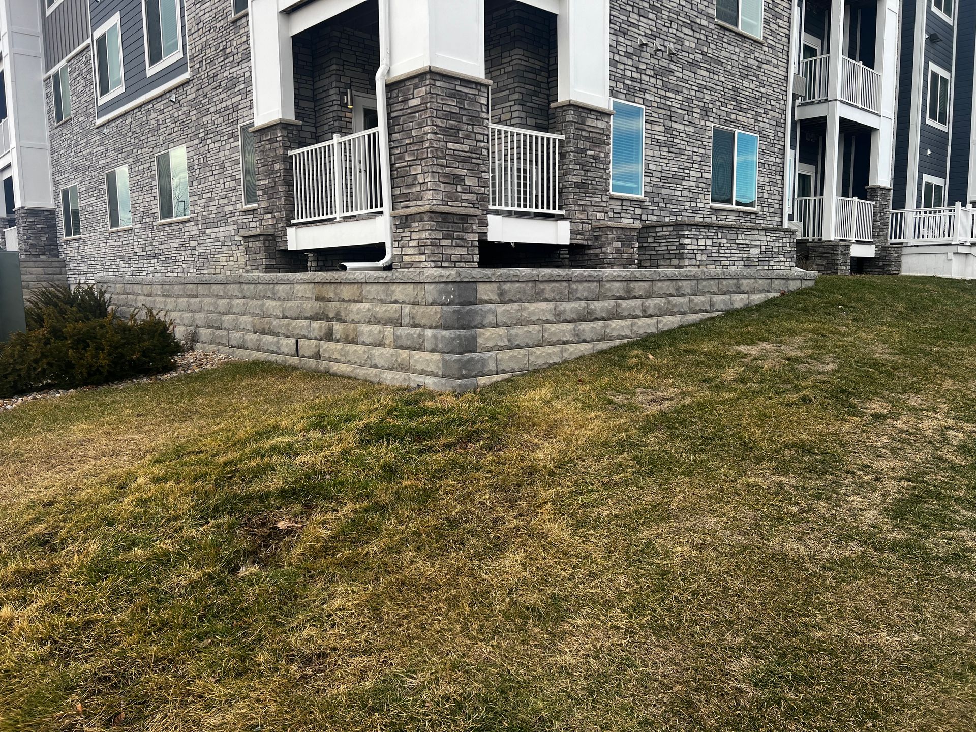 Gray brick retaining wall in front of a multi-story building with a grassy area.