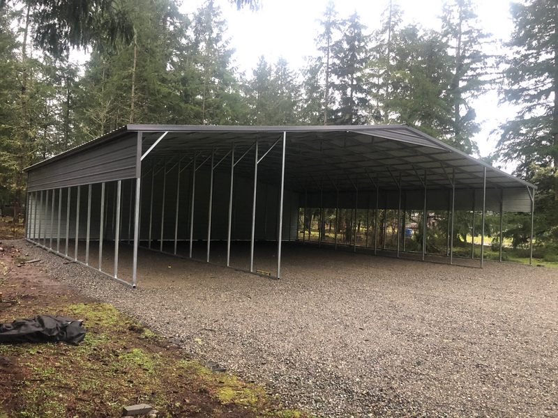 A large metal carport is sitting on top of a gravel driveway in the middle of a forest.