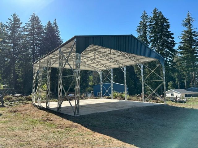 Steel-framed barn-like structure with a concrete floor, green roof, set in a grassy area with trees under a blue sky.
