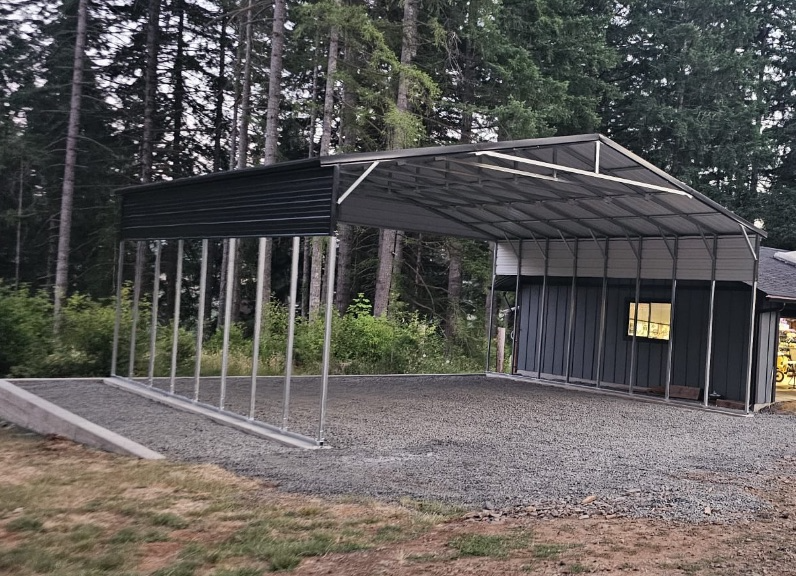 Metal carport next to a small gray building with gravel ground, set in a wooded area.