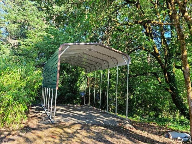 A carport is sitting on the side of a dirt road surrounded by trees.