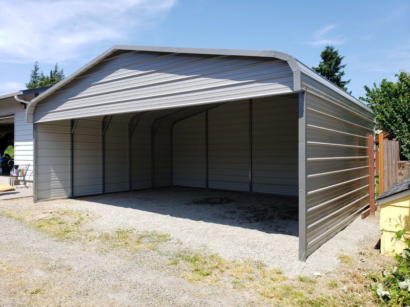 A metal garage is sitting on top of a gravel driveway next to a house.