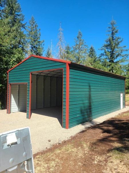 A green metal garage with a red trim is sitting on top of a gravel driveway.