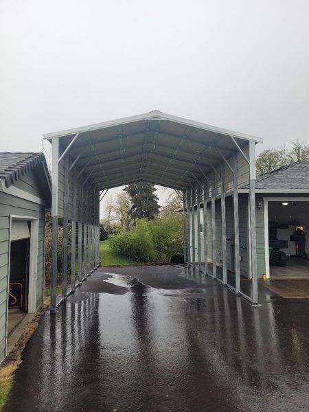 A carport is sitting in the middle of a driveway next to a garage.