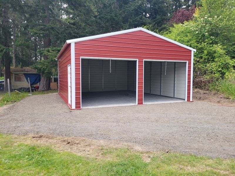 A red garage with two doors is sitting in the middle of a gravel driveway.