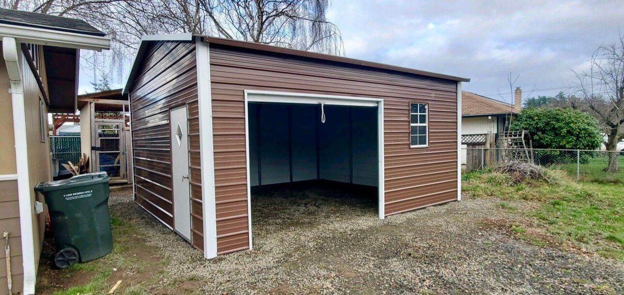 A metal garage with a door open is in the backyard of a house.
