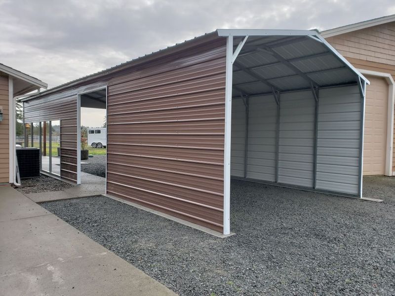 A brown and white metal garage with a covered area in front of a building.