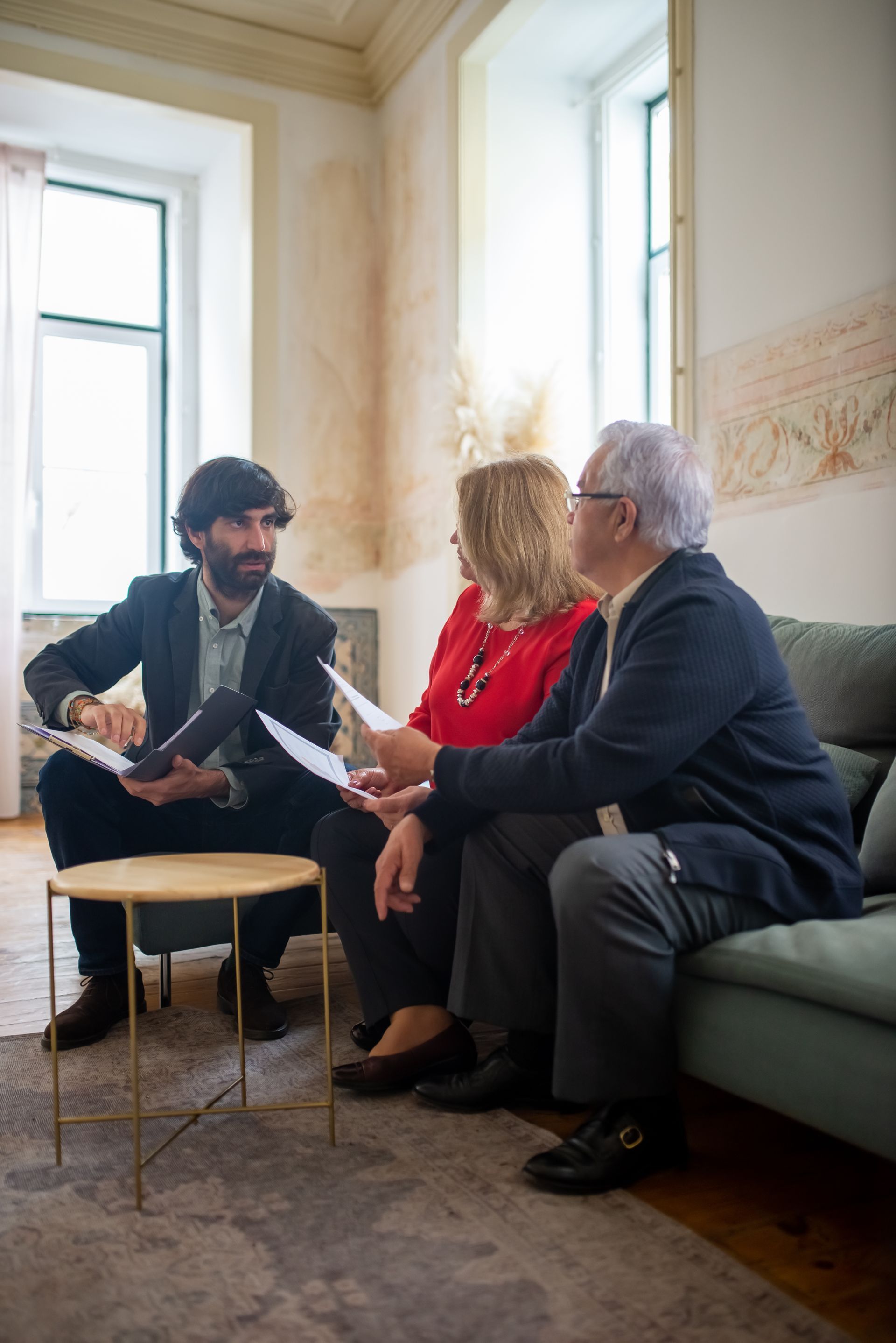 Three people in a bright room discuss papers on a green sofa.
