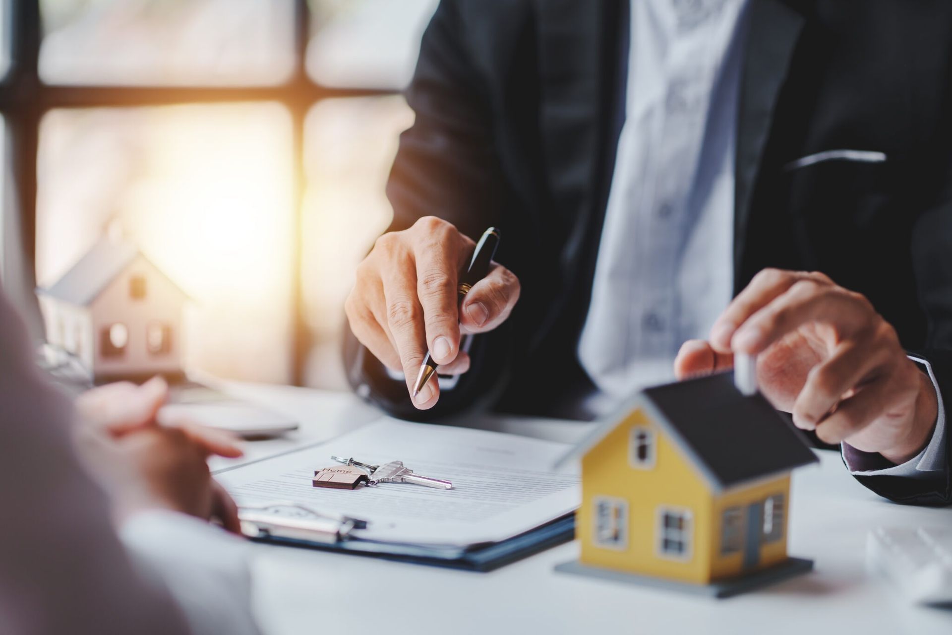 Businessperson signing paperwork beside a yellow model house with keys on a desk
