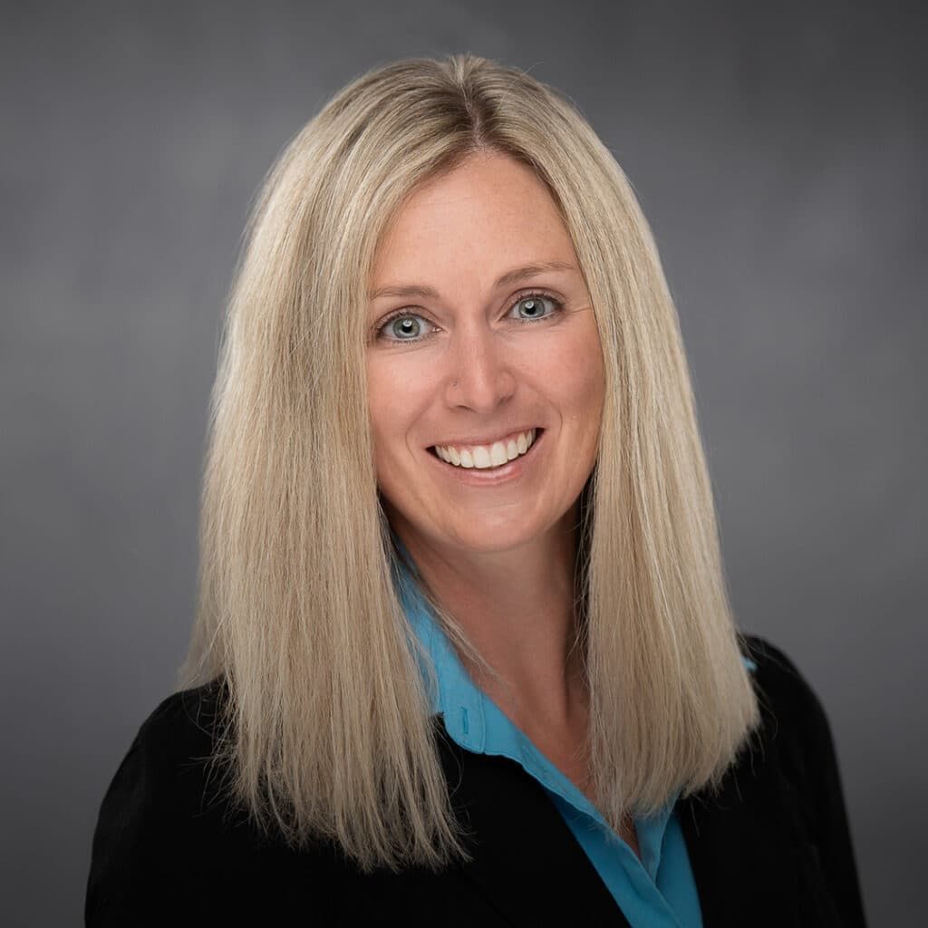 Professional headshot of a smiling woman with long blonde hair wearing a black blazer and blue blouse