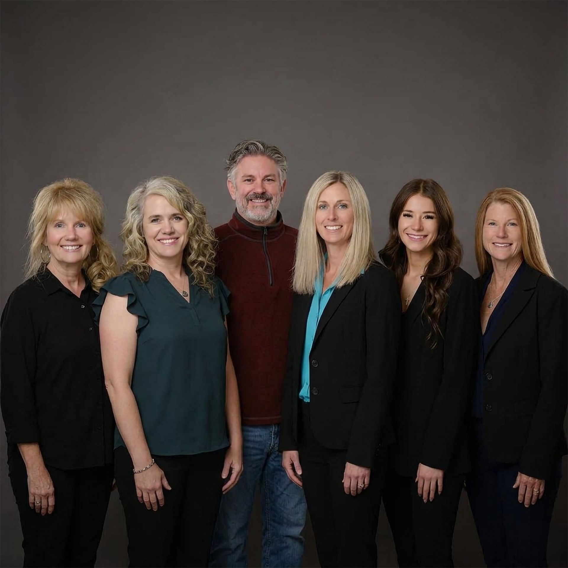 Six people pose together against a gray studio backdrop, dressed in black and dark green clothing.