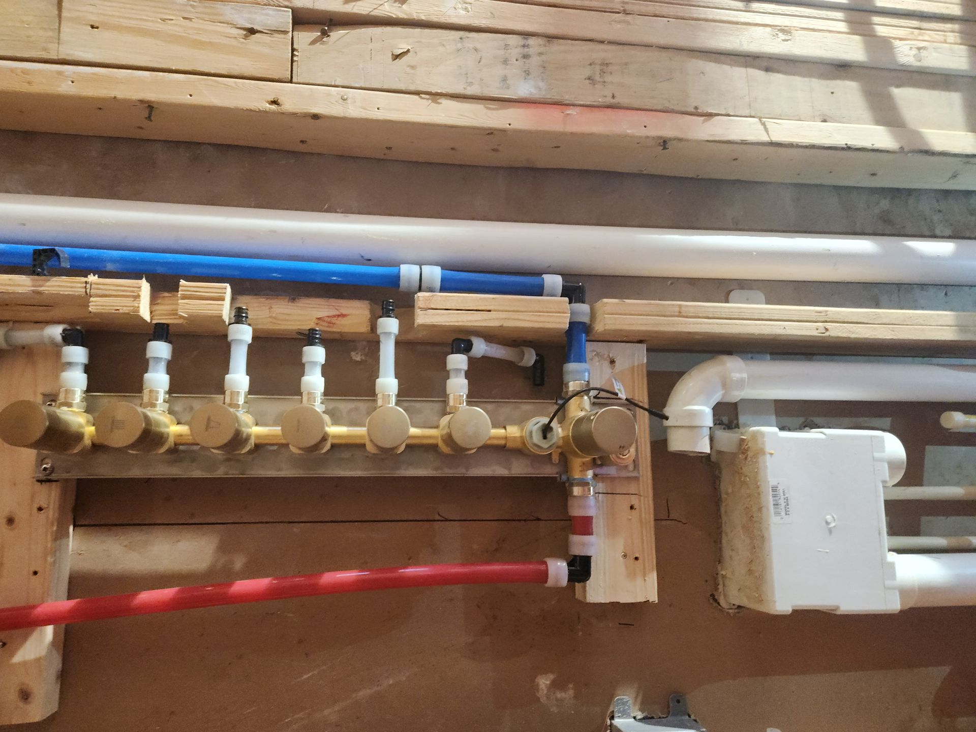 Plumbing manifold with blue and red pipes, brass valves, and a white control box in a wooden wall cavity