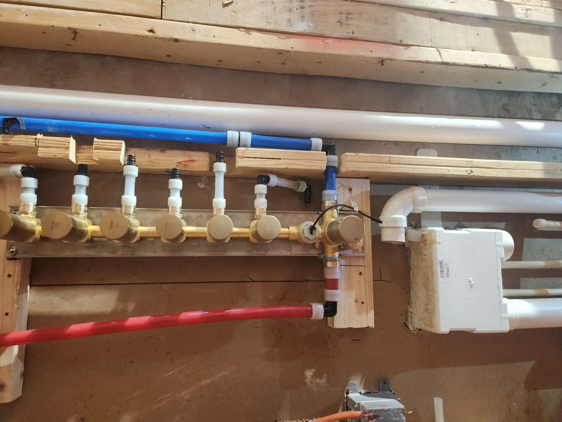 Plumbing manifold with multiple white valves, blue and red pipes, and a white electrical box on a wall.