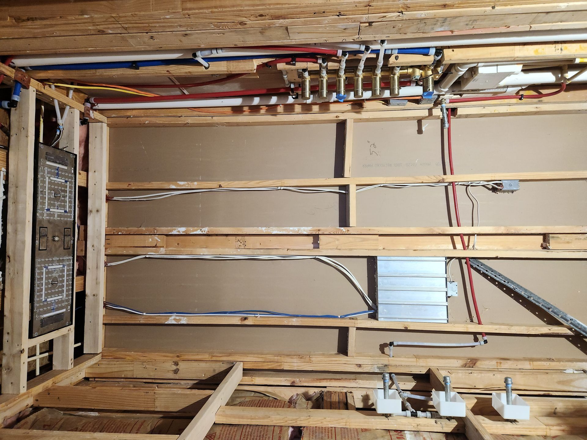 Exposed wood wall framing with wiring, pipes, and an electrical panel in a basement under construction