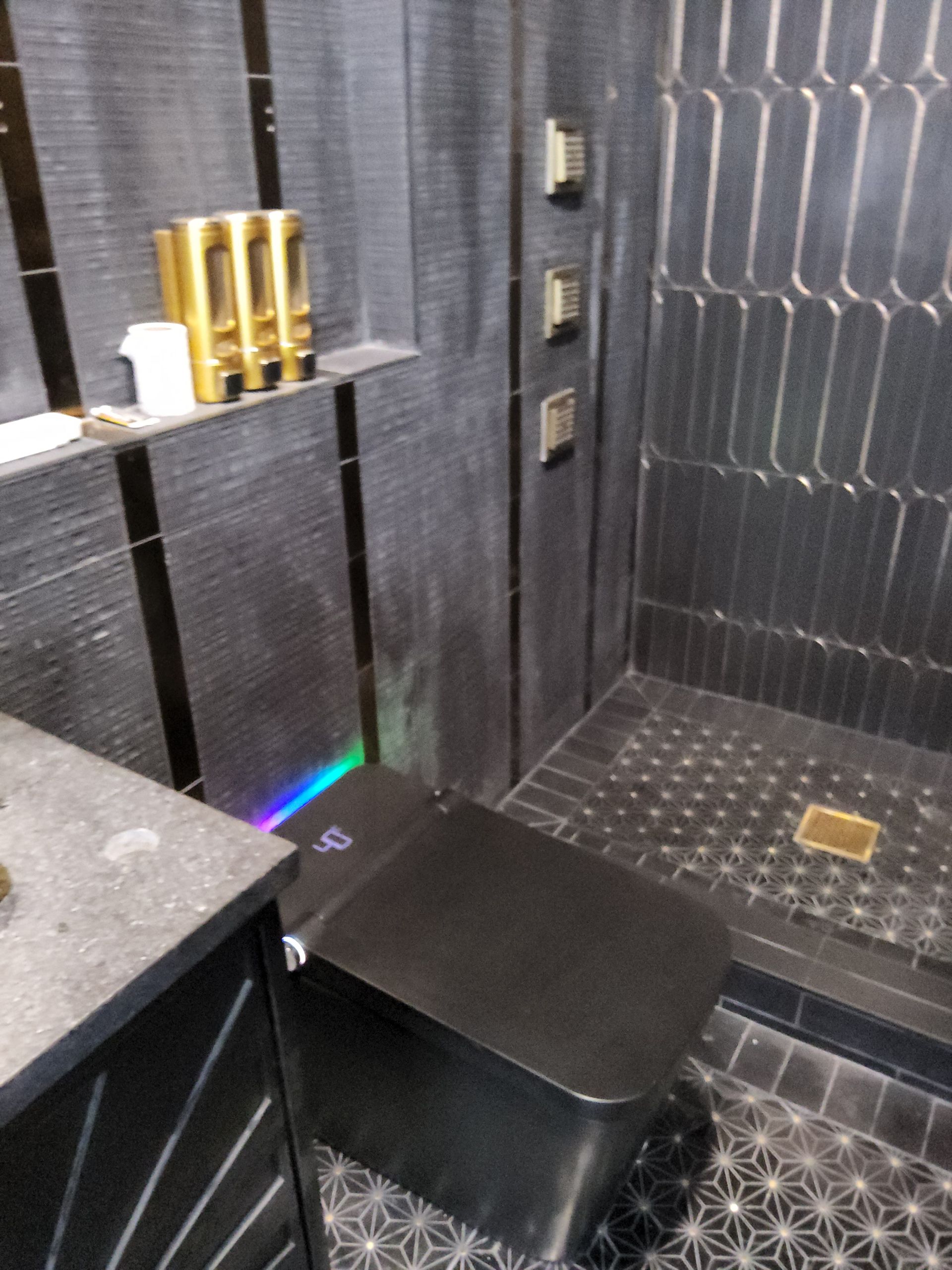 Modern black-tiled bathroom with a wall-mounted toilet and a small shelf holding gold bottles.
