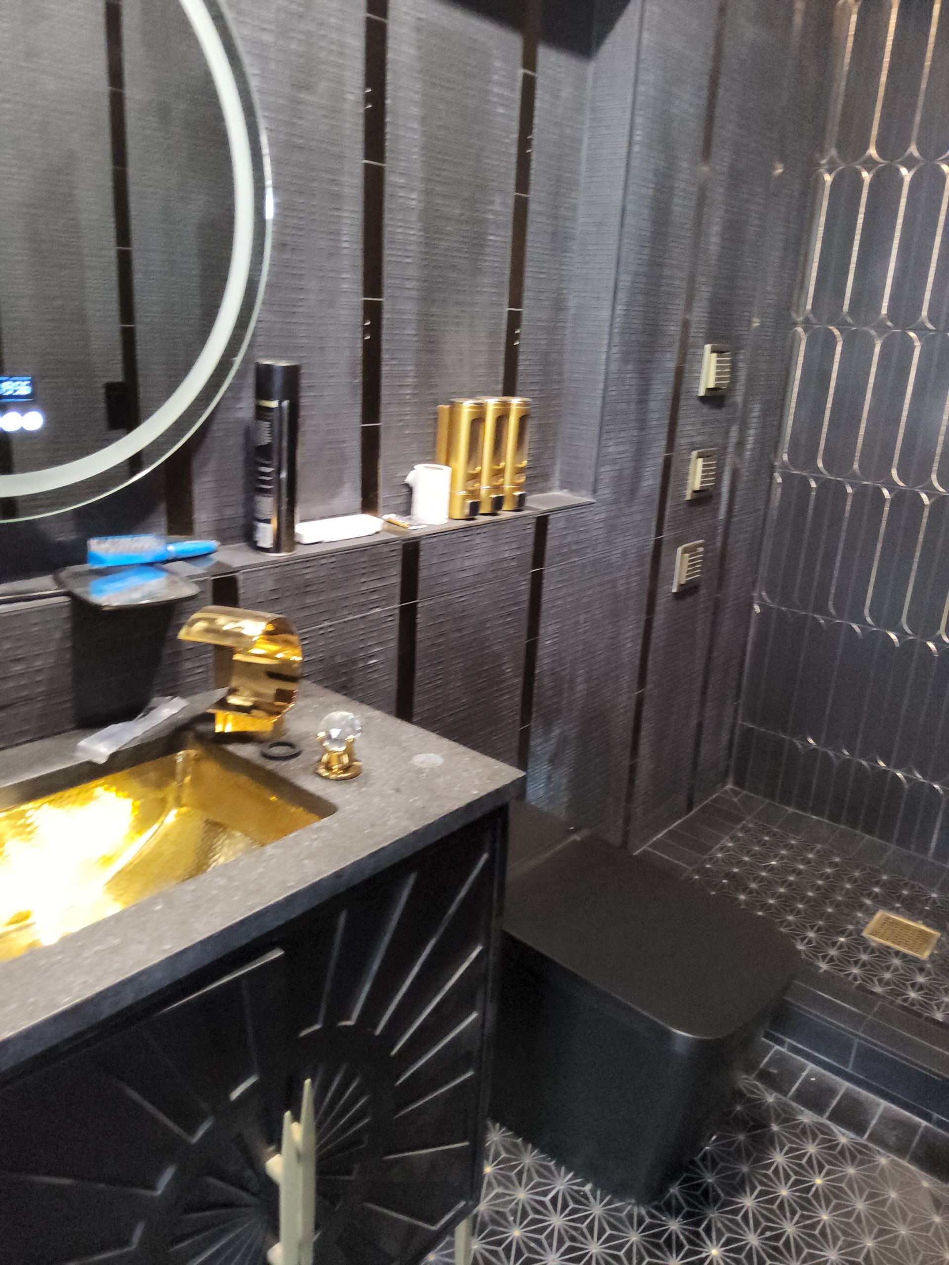 Modern bathroom with black-tiled walls, gold fixtures, and a round mirror above a black sink.