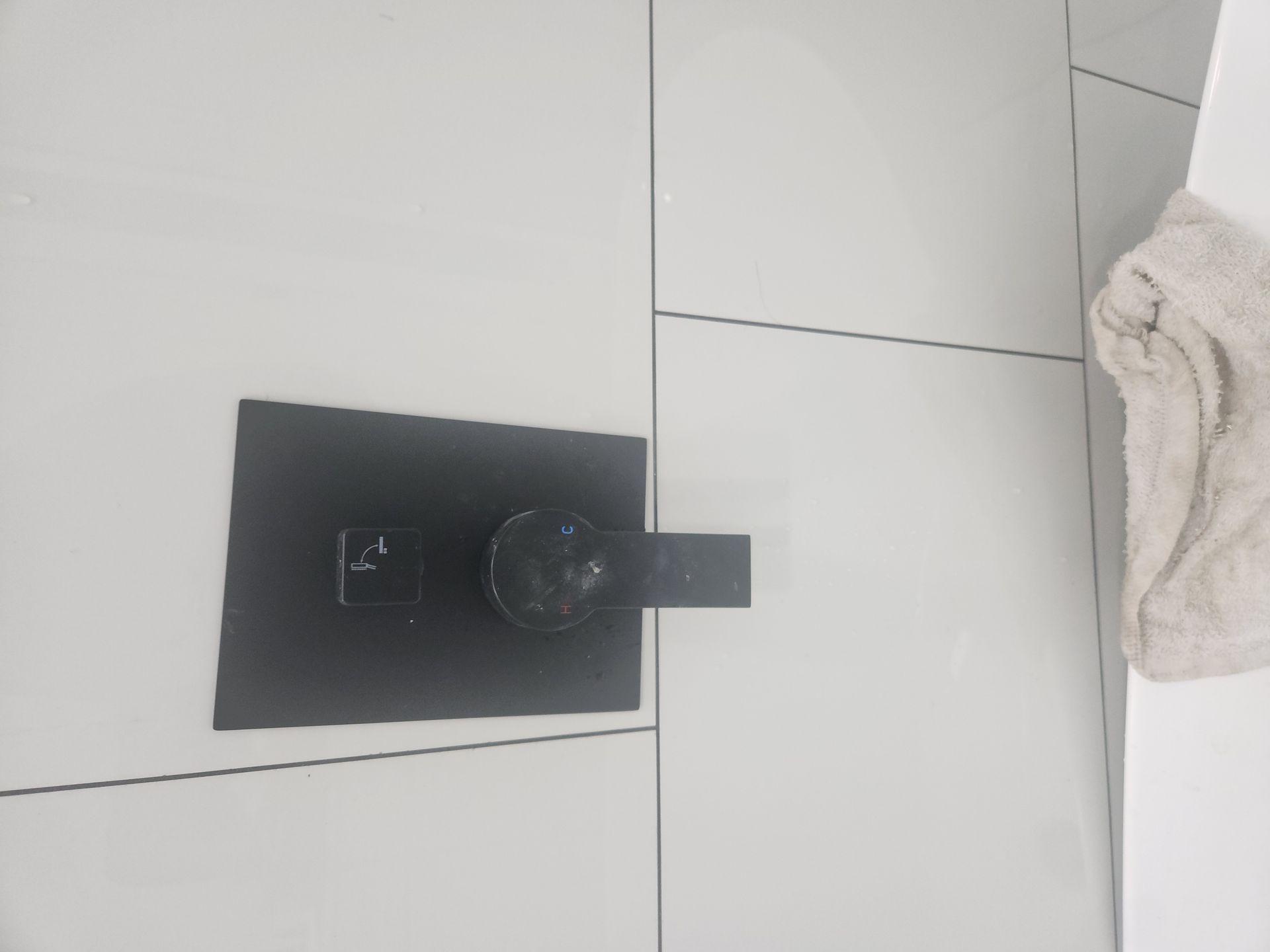 Black wall-mounted control panel on white tiled floor, with a small round knob and nearby cloth.